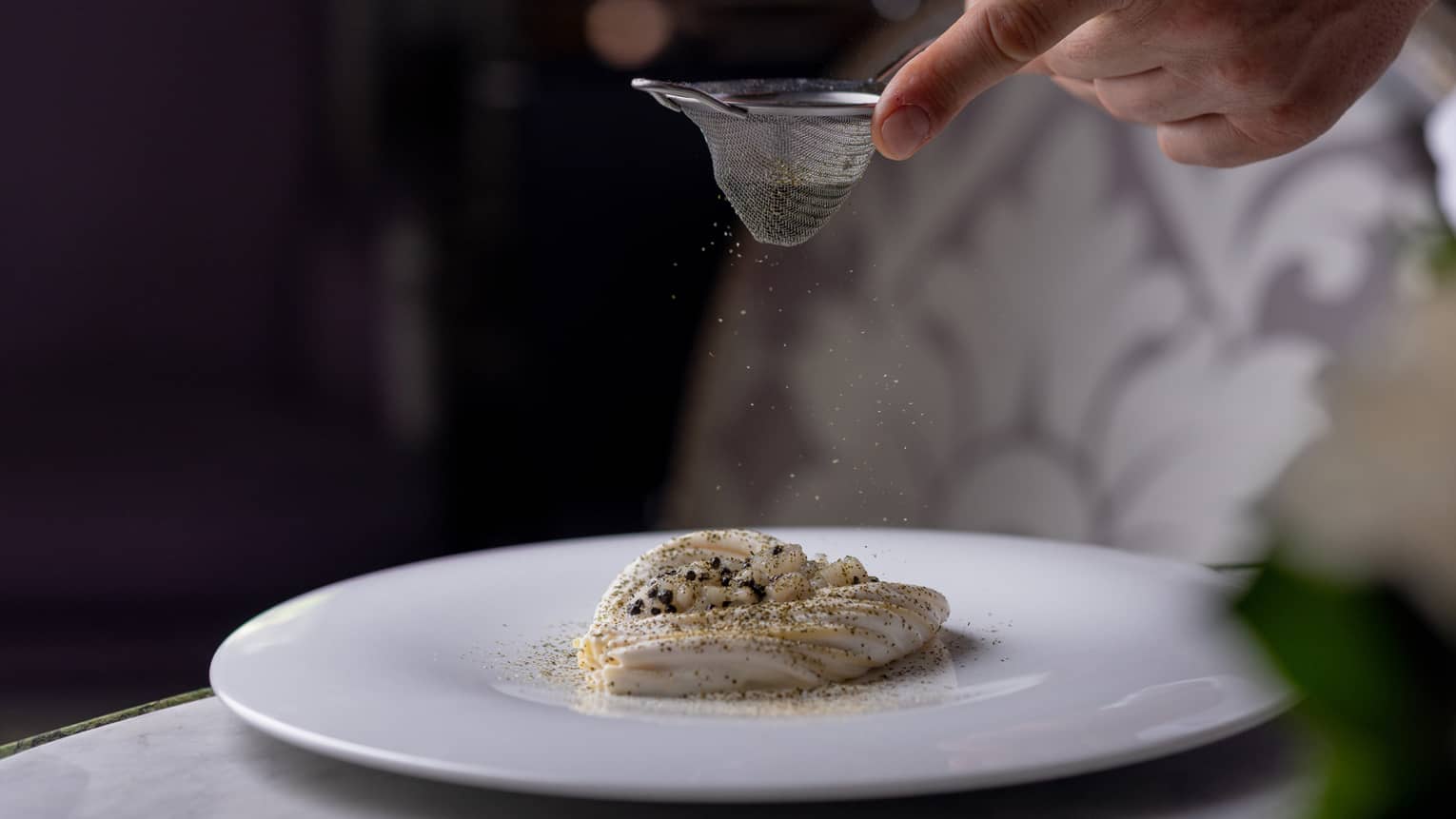 A person sifts a topping onto a food item on a round white plate.