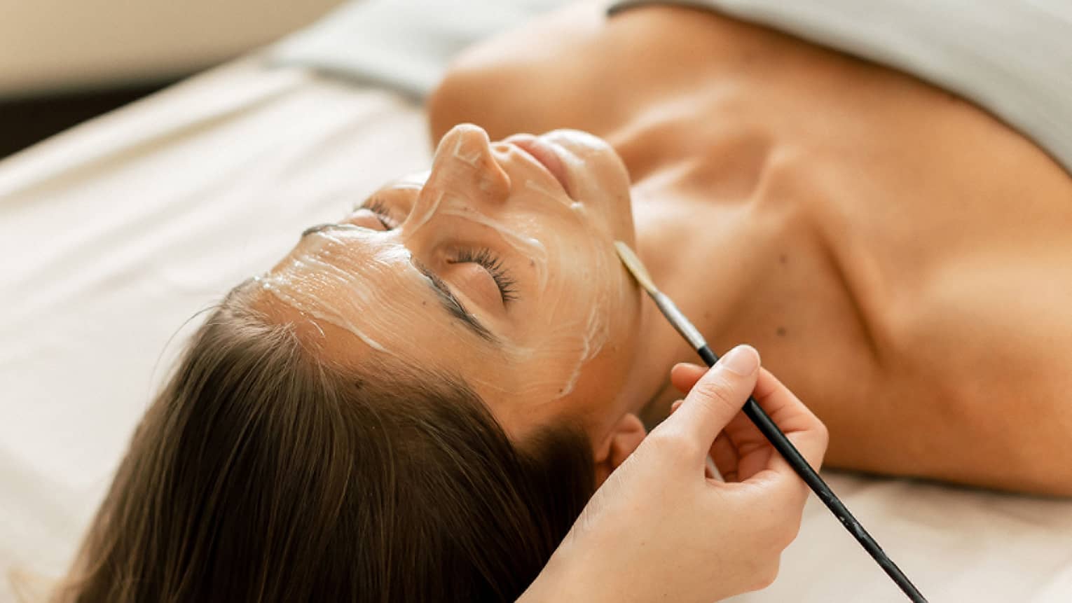 A woman lying on a spa table with eyes closed while a face mask is applied with a small brush