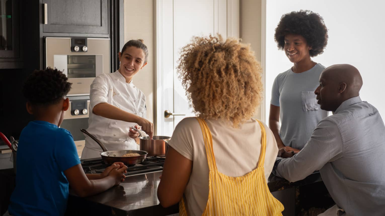 A family of four gathers around a kitchen-island cooktop watching a smiling chef prepare food in copper cookware.