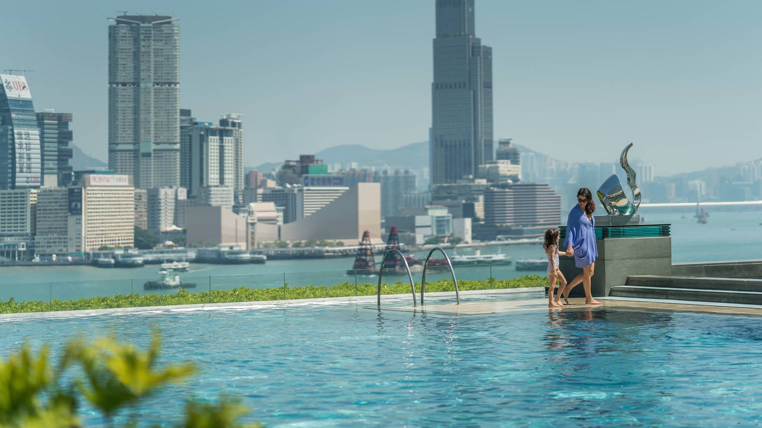 A person and child stand at the edge of a rooftop infinity pool, overlooking the harbour. The clear sky and cityscape create a serene backdrop.