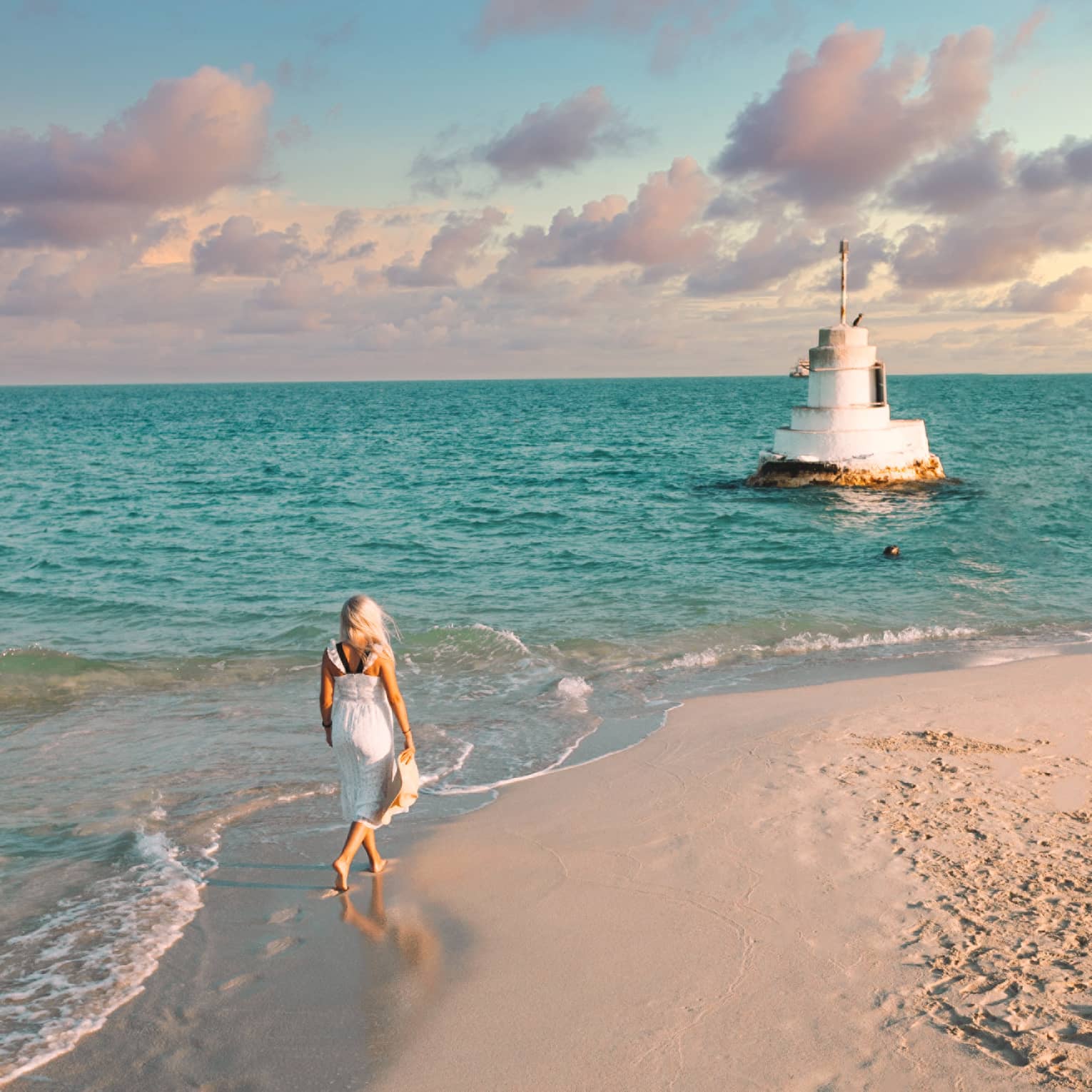 A woman walks along the shoreline towards a stone structure in the sea.