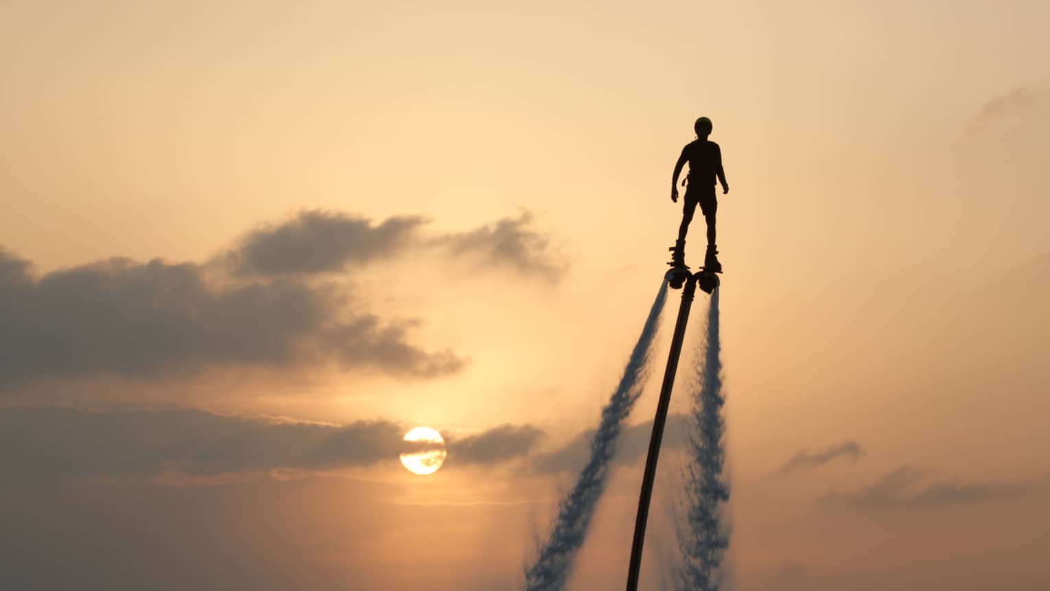 Silhouette of person soaring high above ocean on jet pack, water trails below, against sunset