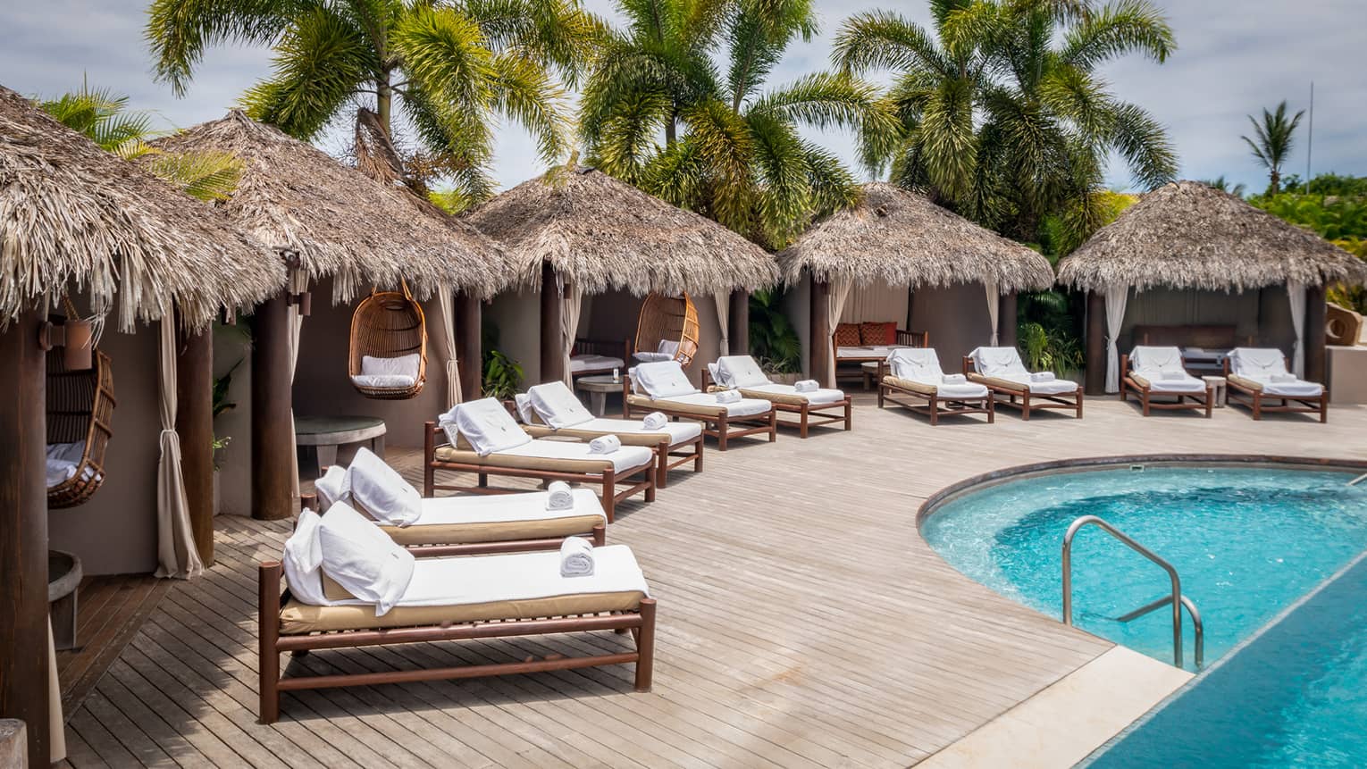 Grass-topped poolside cabanas with row of lounge chairs in front, palm trees in distance