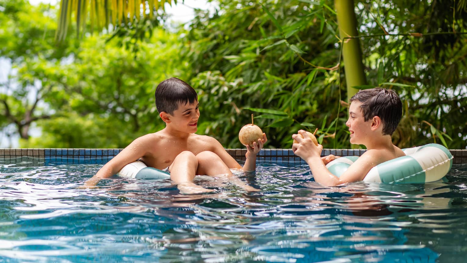 Two children float on green-and-white-striped inner tubes in a pool surrounded by lush greenery