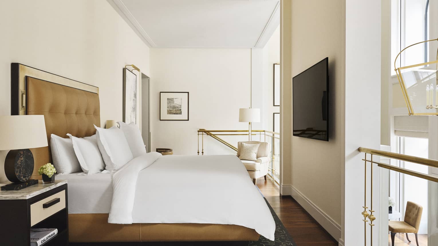 Luxury hotel loft suite bedroom with a king bed with plush headboard, wall-mounted TV, armchair, bedside table with lamp and gold railing