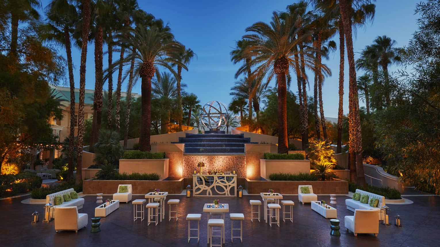 Palm trees over stone fountain, terrace seating area with lights at dusk