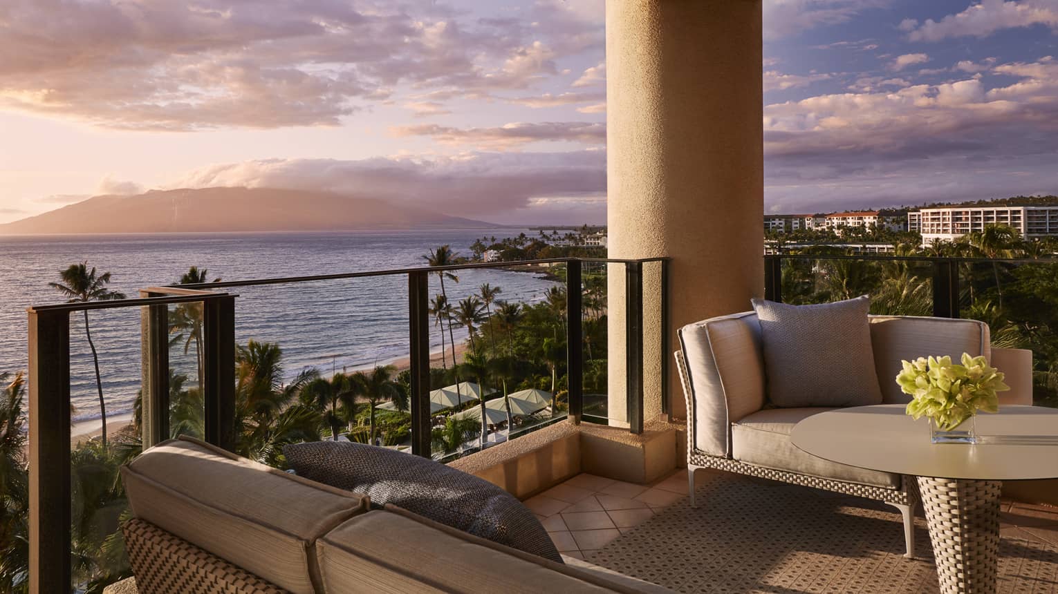 Balcony with cushioned seating and table overlooking beach palm trees and ocean at sunset