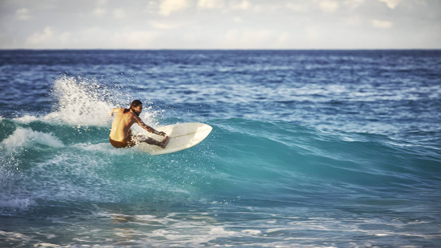 On an expanse of water, a surfer leans back on his board and surfs the face of a wave as white water splashes up behind him.