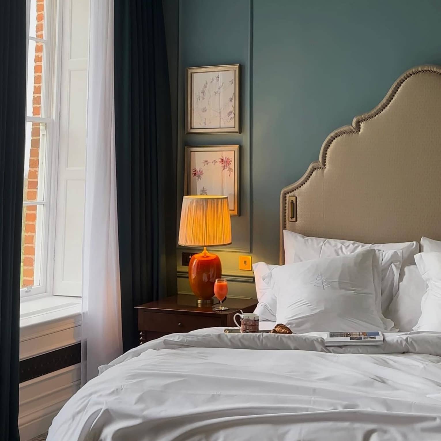 A magazine, mug and pastry sit atop a bed with a large cushioned headboard next to a window