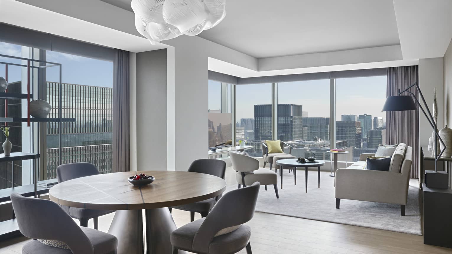 Hotel suite with round dining table with chairs, seating area, modern shelving and floor-to-ceiling windows with panoramic city views