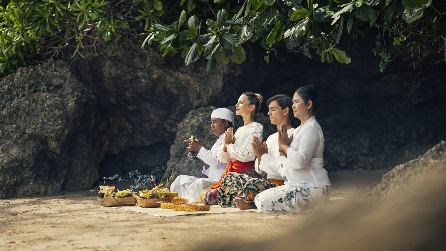 Four adults sit on the ground beside a rock face with their eyes closed and hands held together in front of their chests.