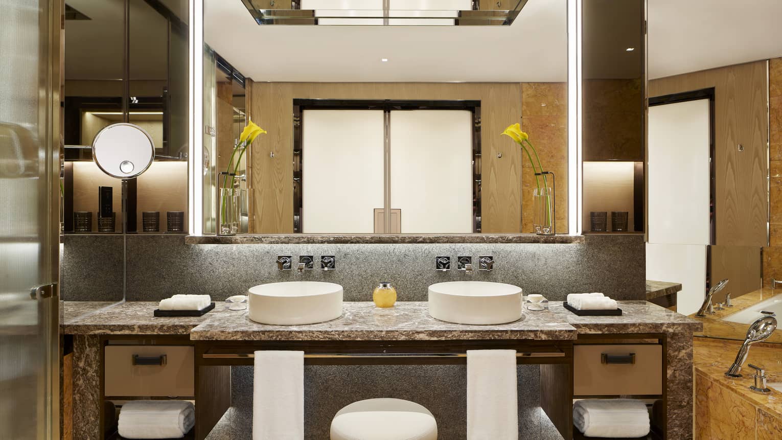 Luxurious bathroom in the Deluxe Peak-View Room with double vanity, marble countertop, tall mirrors, folded towels, a bathtub, and elegant lighting fixtures.