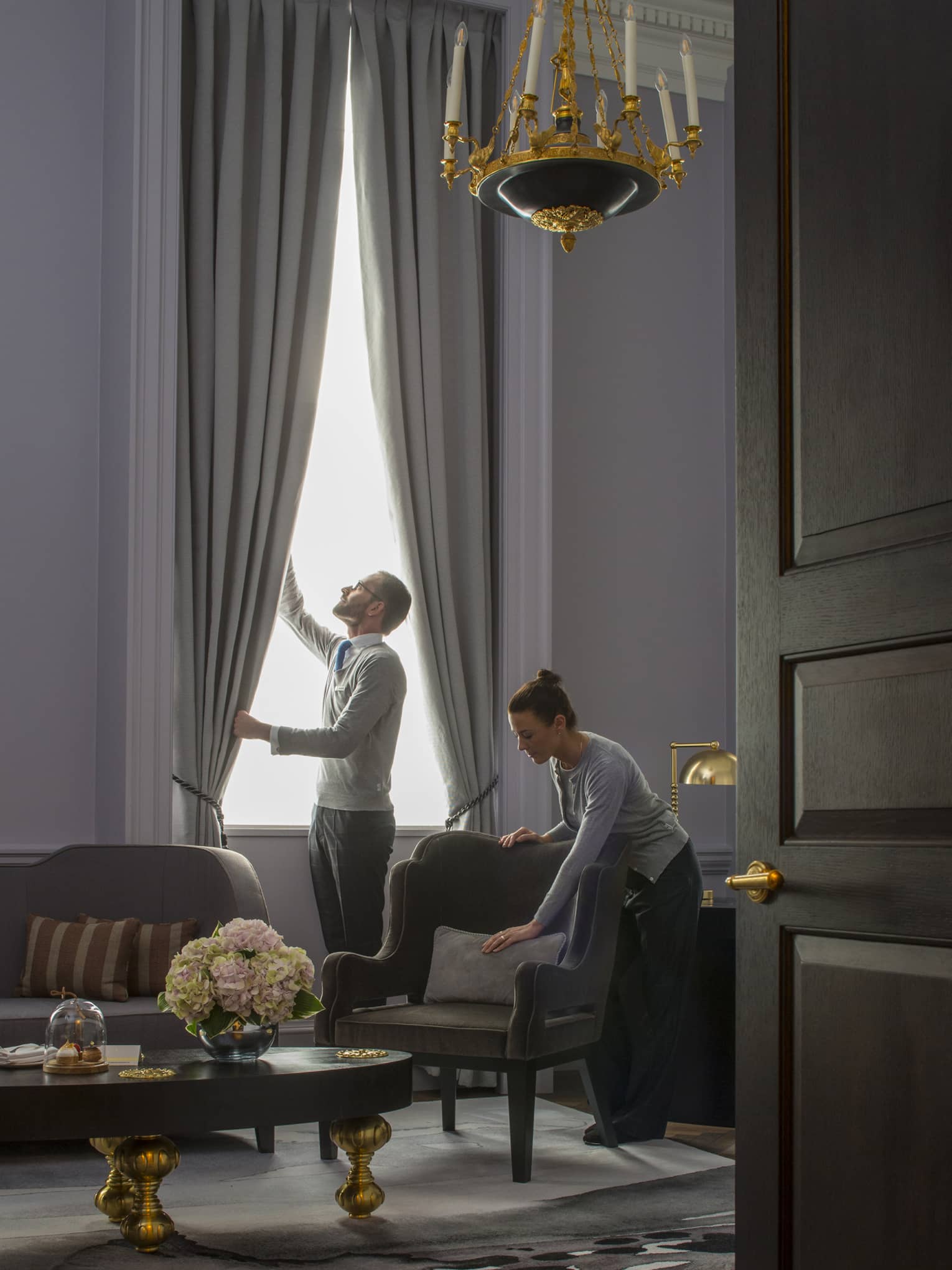 Grand Heritage Suite, man opens drapes as woman smooths pillow on armchair under chandelier