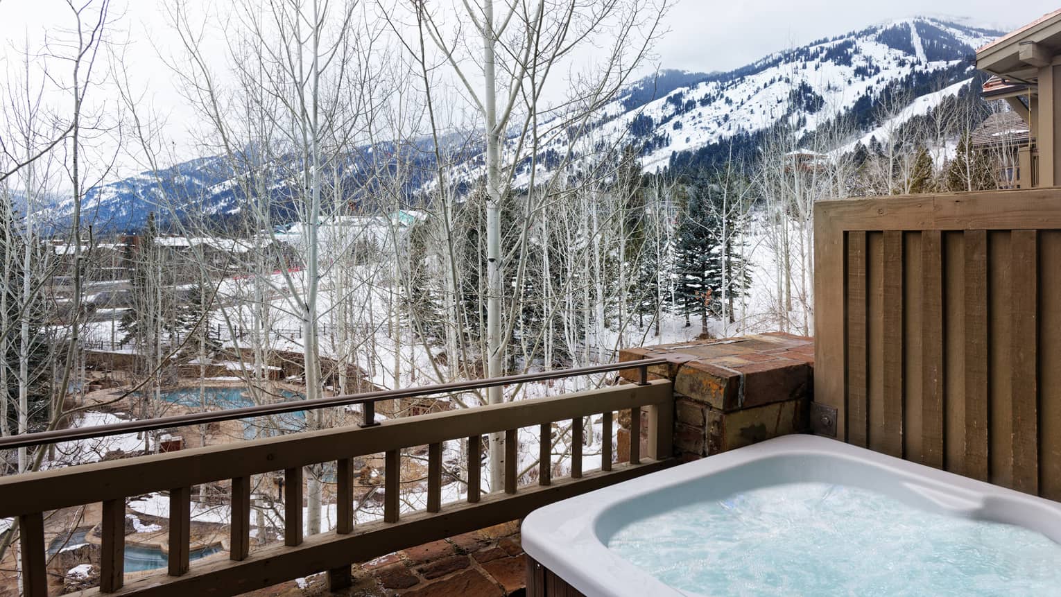 Outdoor hot tub on a private balcony of a luxury resort residence with a view of snow-covered mountains, bare trees and a nearby pool area below