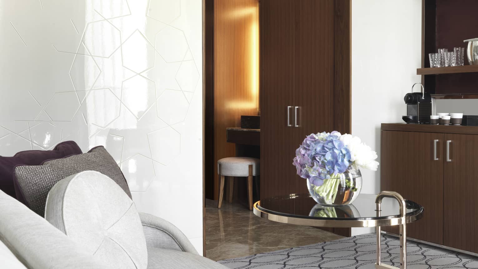 Modern hotel suite with a gray sofa, purple cushions, a glass table with a vase of blue and white hydrangeas and a wooden minibar in the background