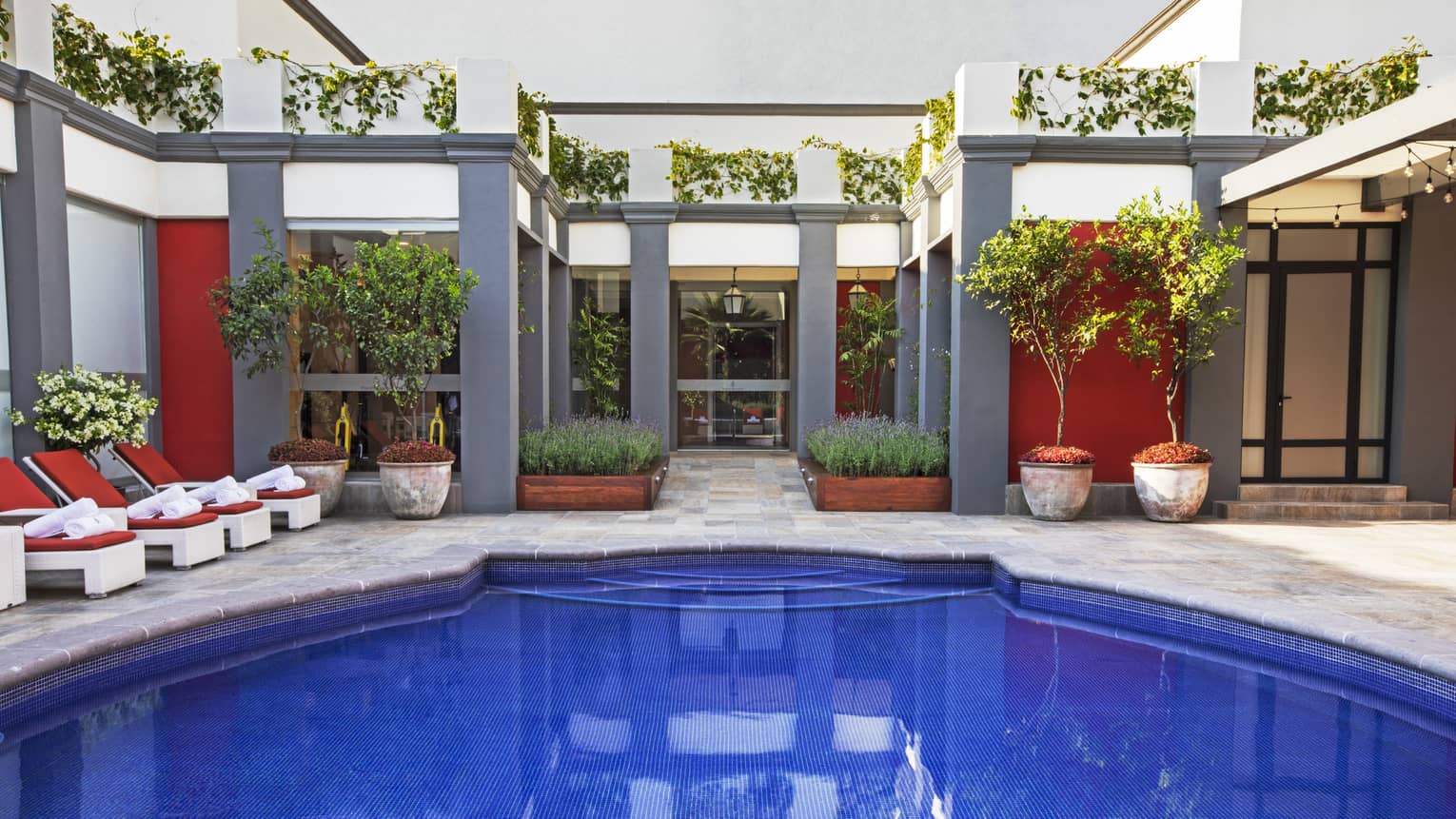 A brightly lit outdoor pool with deep blue water with red lounge chairs, plants, and open space around it.