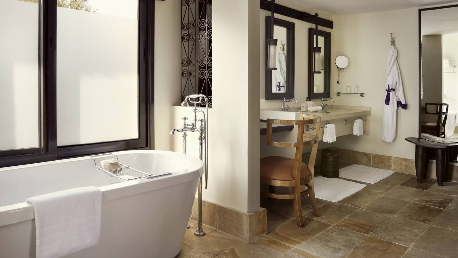 Bathroom with deep soaking tub against window and double vanity under rustic mirrors with brown desk chair, all on slate flooring