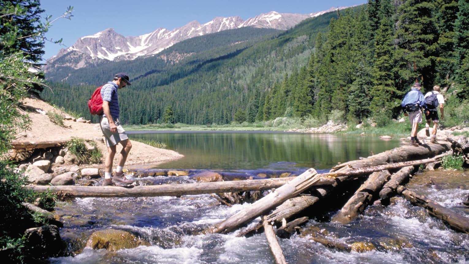 Two young hikers ahead of an adult walking on fallen logs over river rapids against a majestic, forested mountain backdrop.