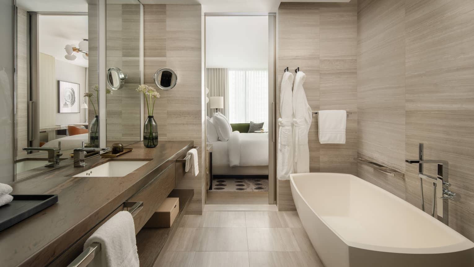 Luxury hotel bathroom with a freestanding tub, spacious vanity, large mirror, and two white bathrobes hanging beside the door