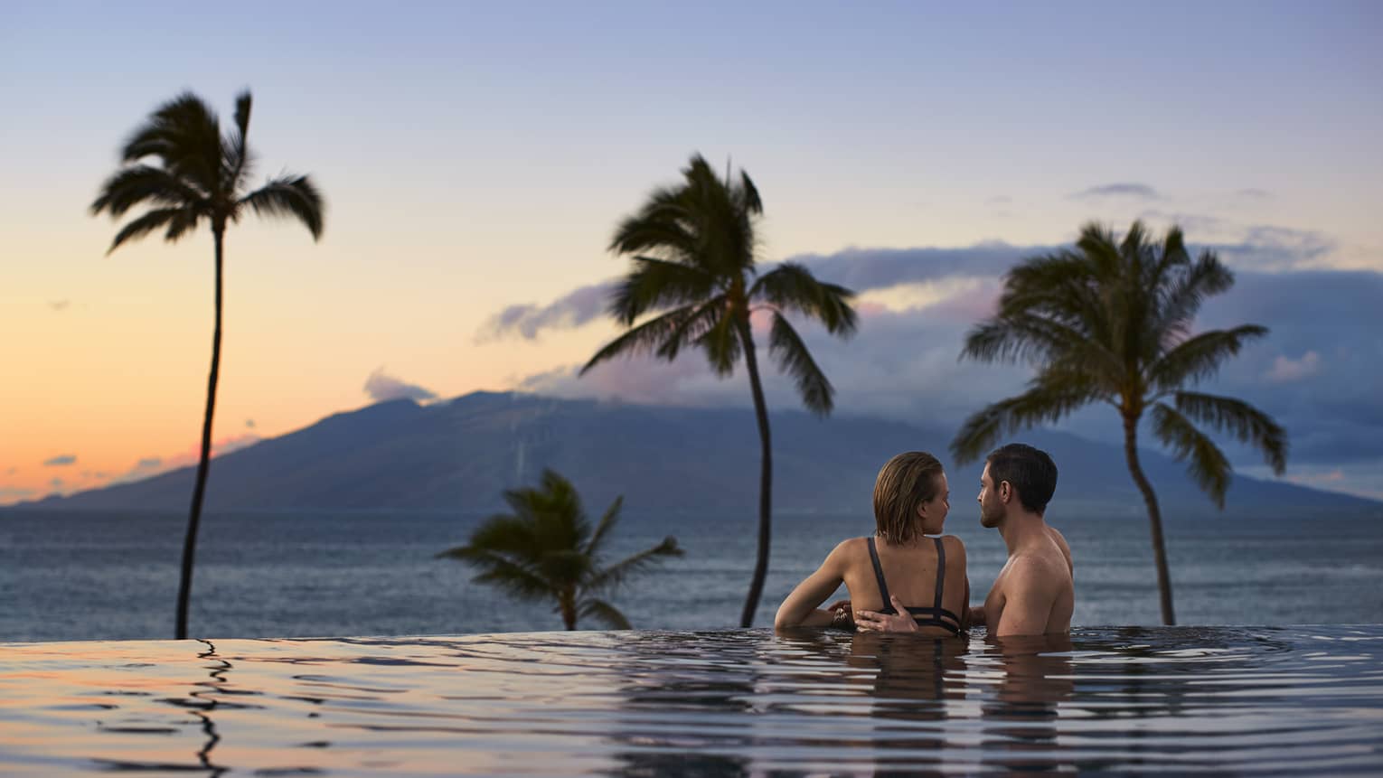 Couple embracing in an infinity pool at sunset, overlooking the ocean and distant mountains with palm trees