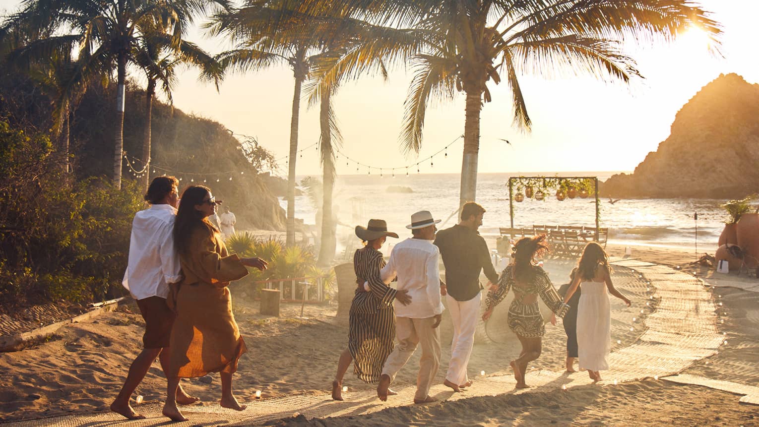 A group of adults walks down a palm-lined beach path towards a table by the ocean as the sun sets behind a rocky outcrop.