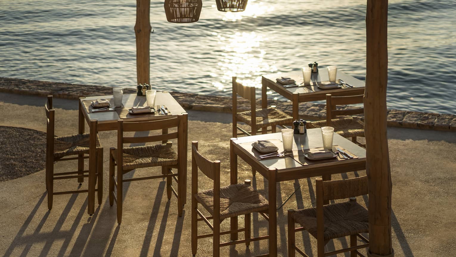 Outdoor taverna next to ocean with three wooden tables and chairs, three wooden lamps