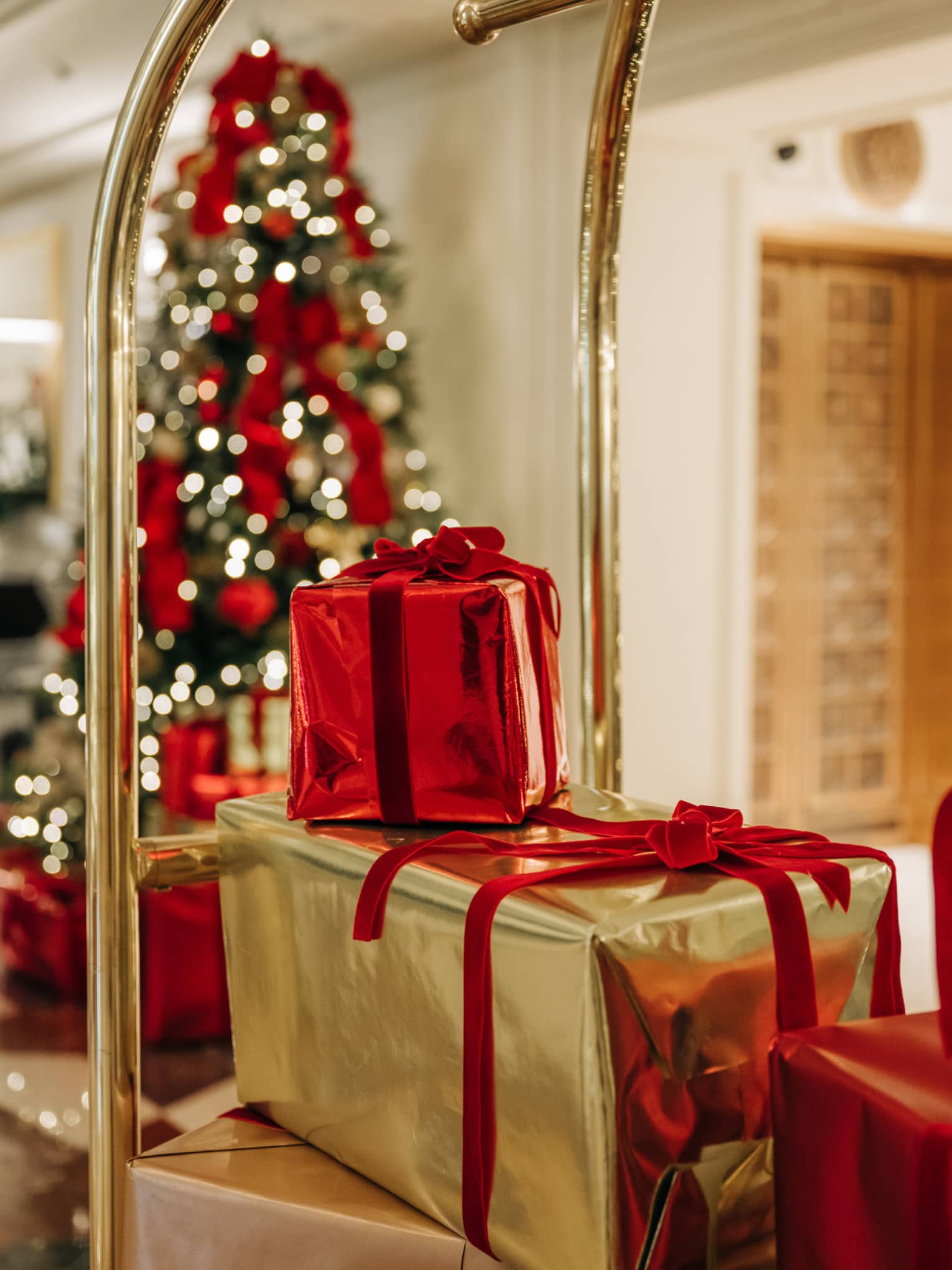 Shiny red and gold present boxes being delivered to a hotel room on a gold luggage cart pass by a Christmas tree near an elevator