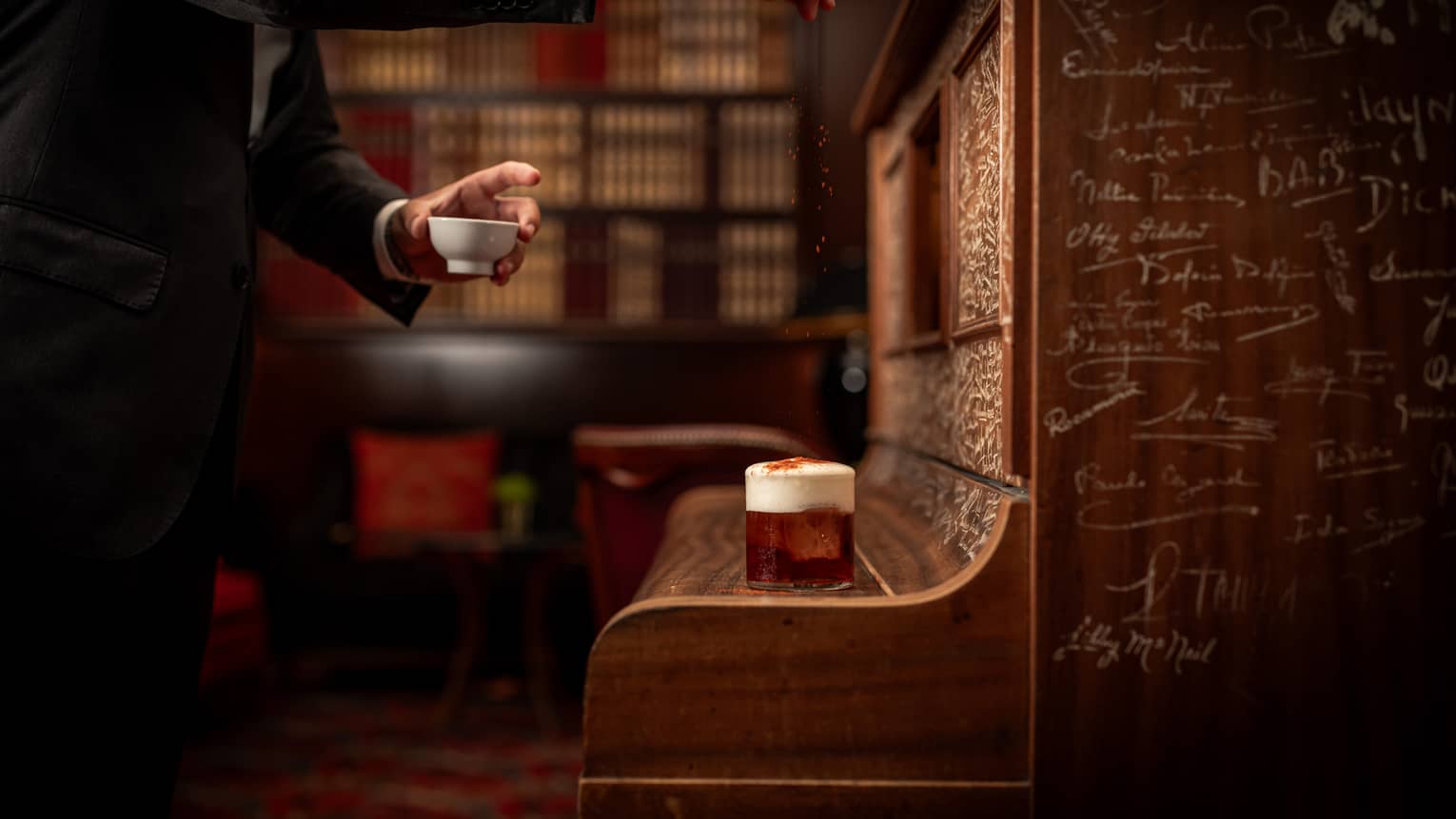 A man dressed in a black suit sprinkles a garnish on top of a cocktail that sits atop a wooden bench