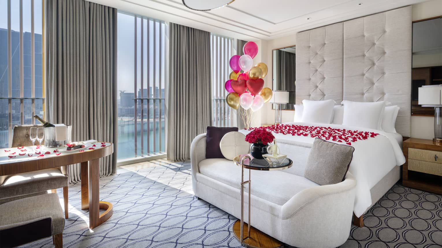 Hotel guest room with a white bed, white setee, desk and wall of floor-to-ceiling windows decorated with red rose petals on the bed and gold, pink and white balloons