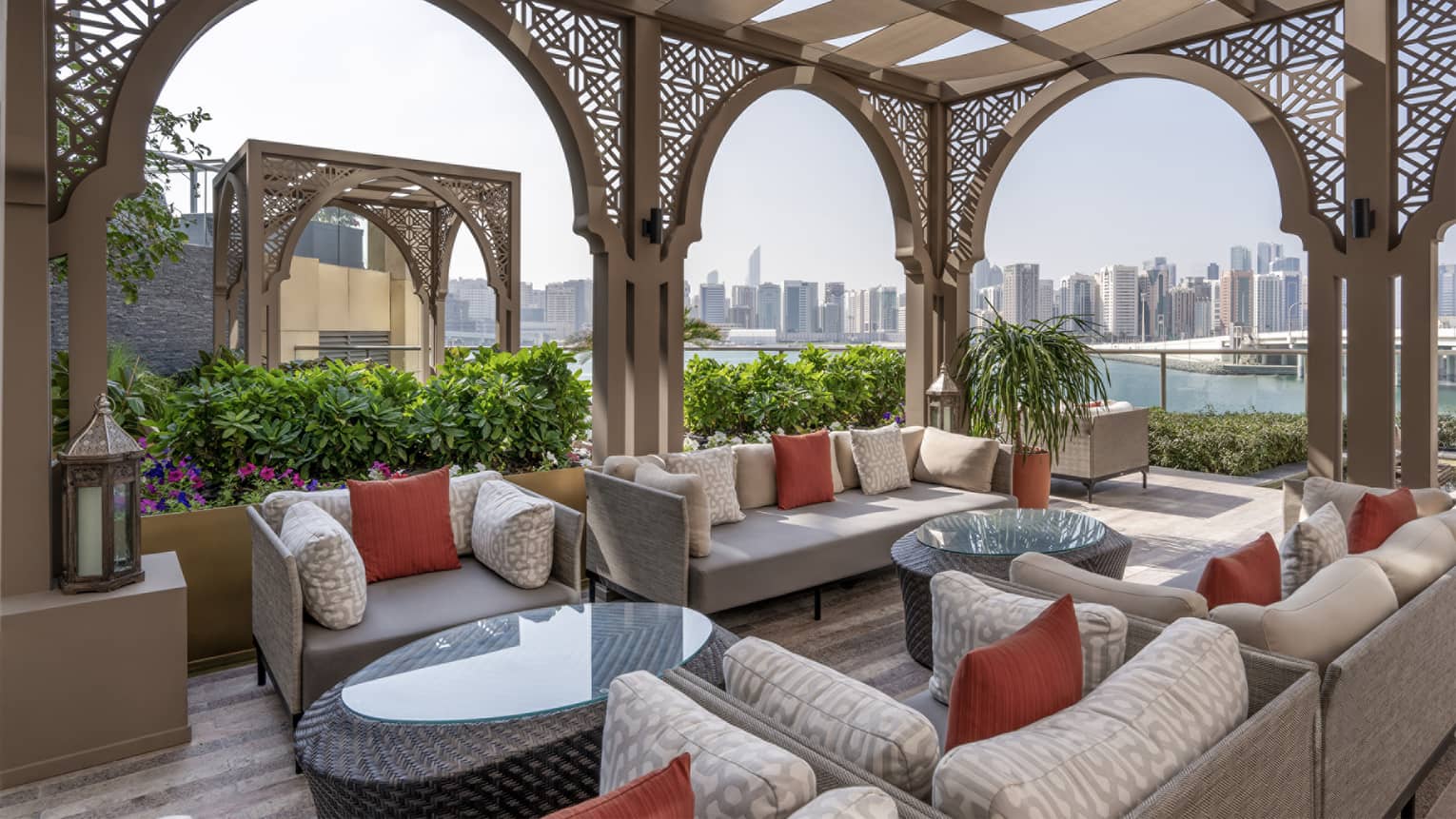 Outdoor terrace with light-grey lounge seating set beneath an Arabian-style pergola with a city skyline seen in the background