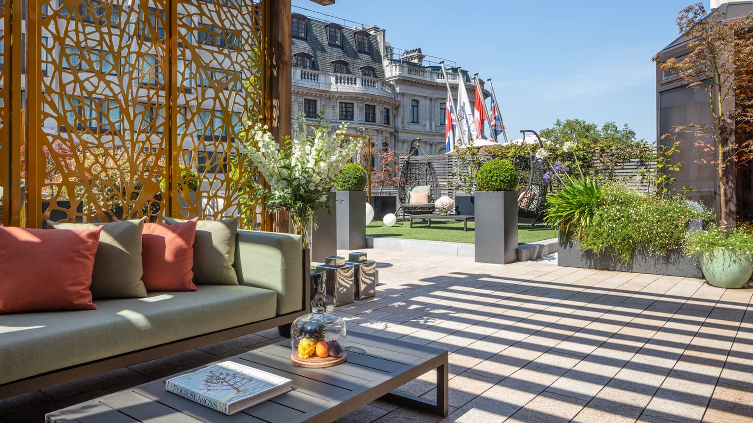 The Terrace Suite at Four Seasons London Park Lane in Mayfair features an expansive and beautifully designed outdoor terrace. The space is adorned with modern outdoor furniture including a sleek sofa with plush cushions and a low coffee table, perfect for relaxation or entertaining. The terrace is surrounded by lush potted plants, sculptural elements, and features decking that adds to the sophisticated ambiance. The view includes the tops of surrounding historic London buildings, creating a luxurious and serene urban garden atmosphere.