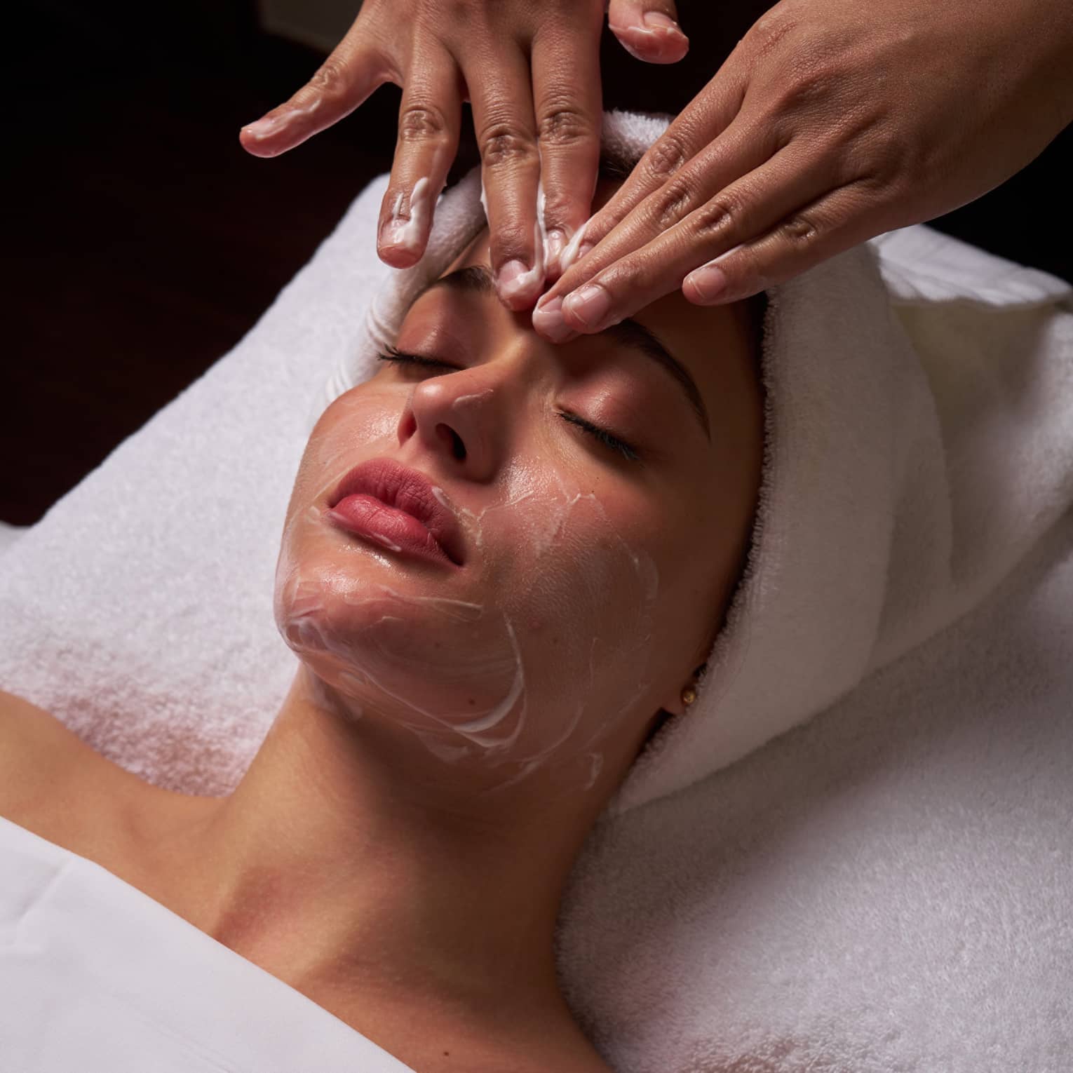 Person lies on massage table covered in a white cloth from shoulders down with hair wrapped in white towel while a pair of hands massages their forehead
