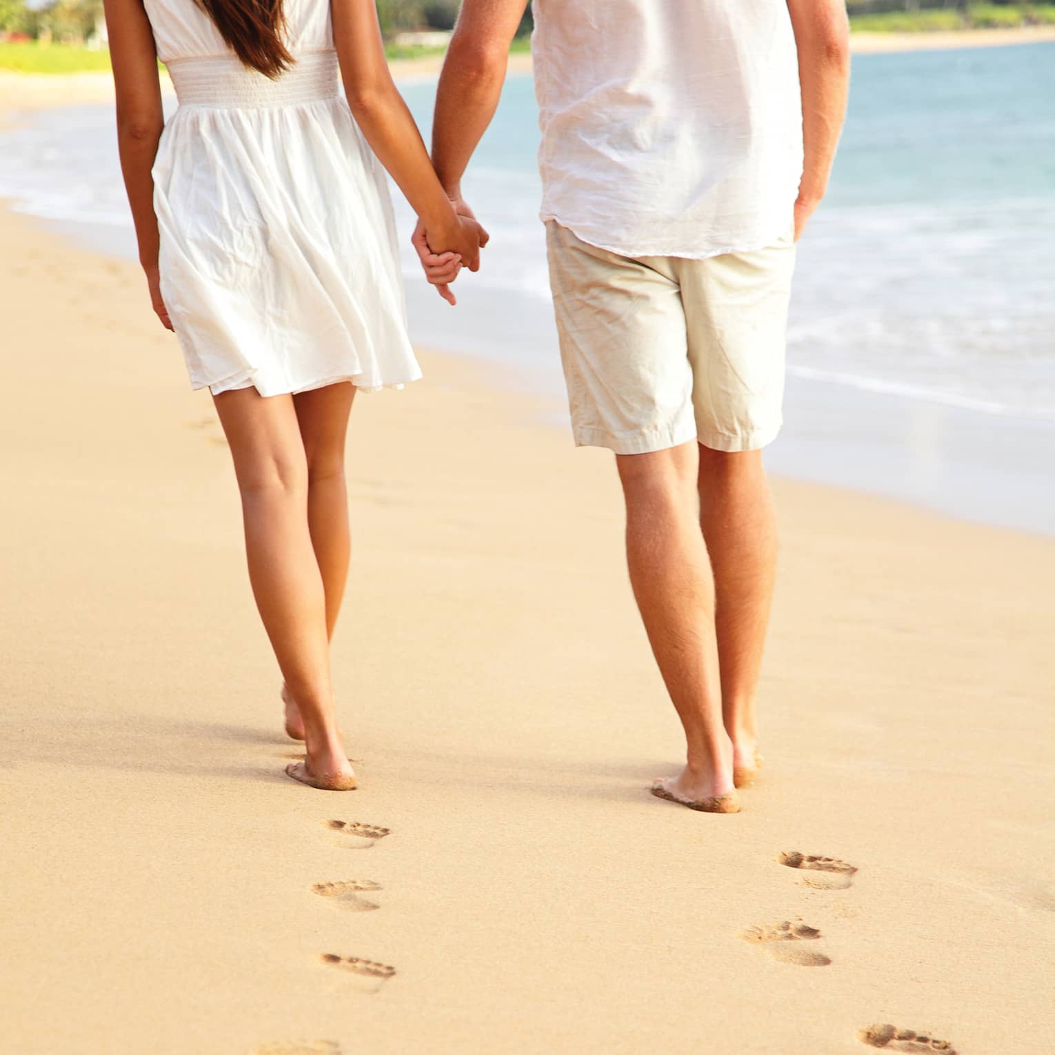 A close-up of a couple holding hands and leaving a trail of footprints as they stroll down a beach near the water’s edge.