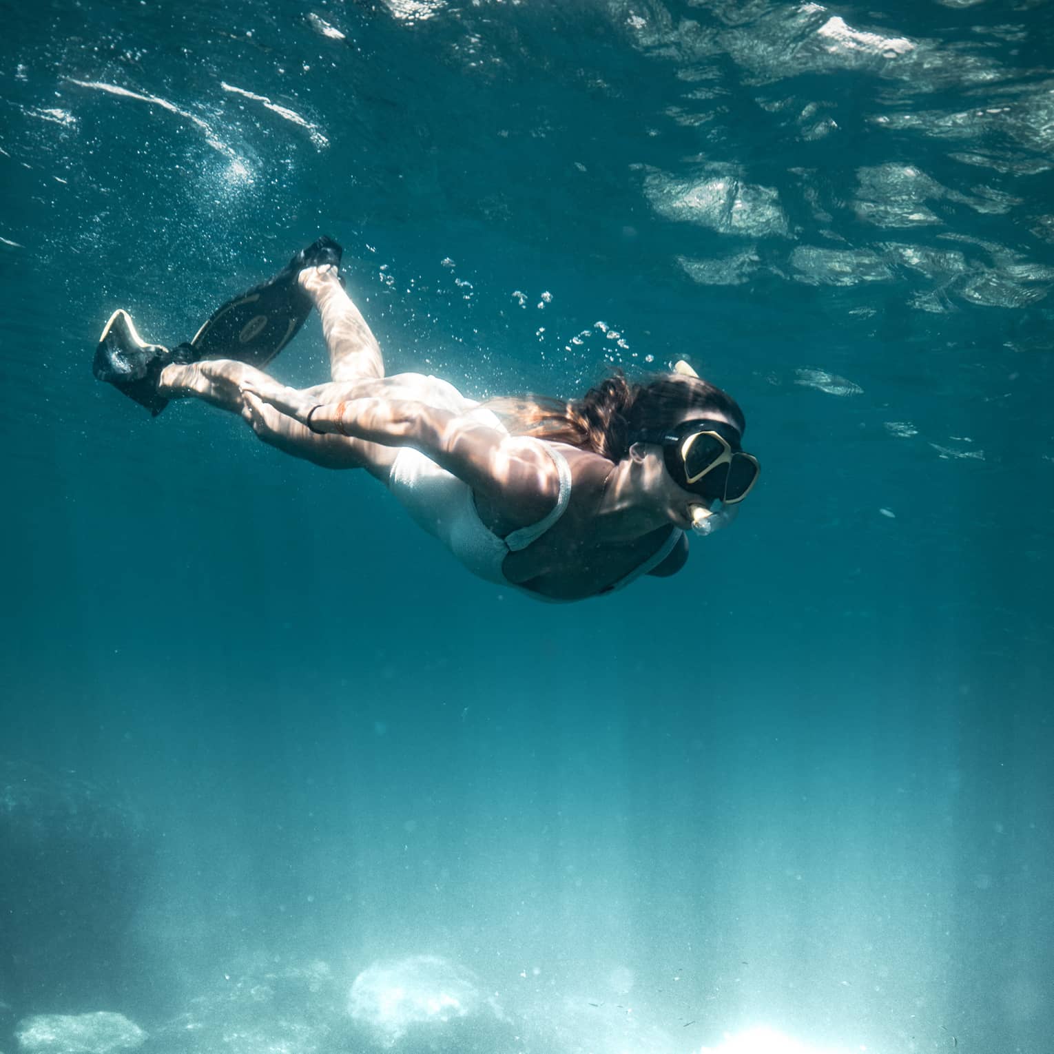 Light filters below the water’s dark blue surface as a diver with fins, goggles and a snorkel kicks toward the seafloor.