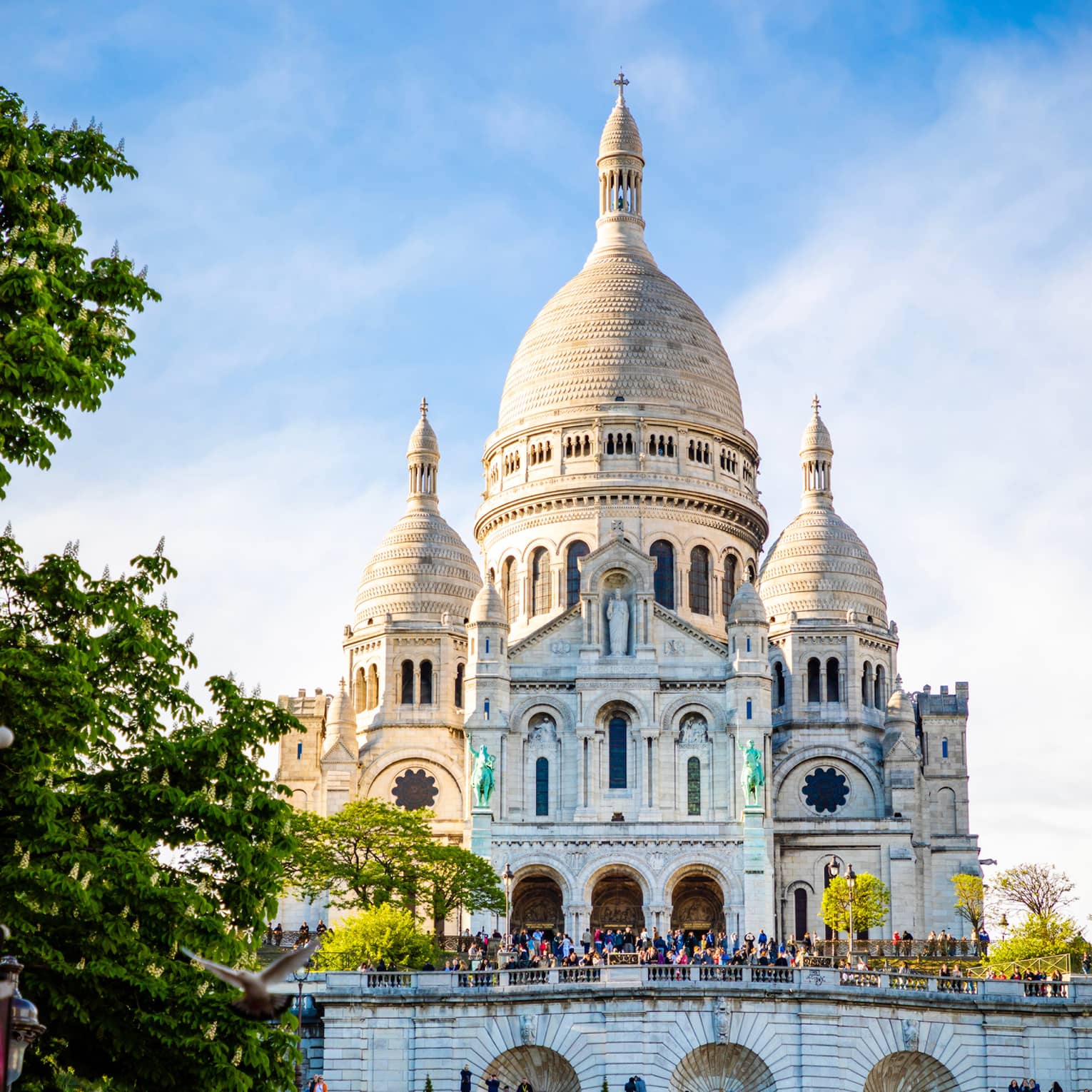 Sacré-Coeur Basilica sits atop a grassy hill under a cloudy blue sky as tourists mill about near the entrance.