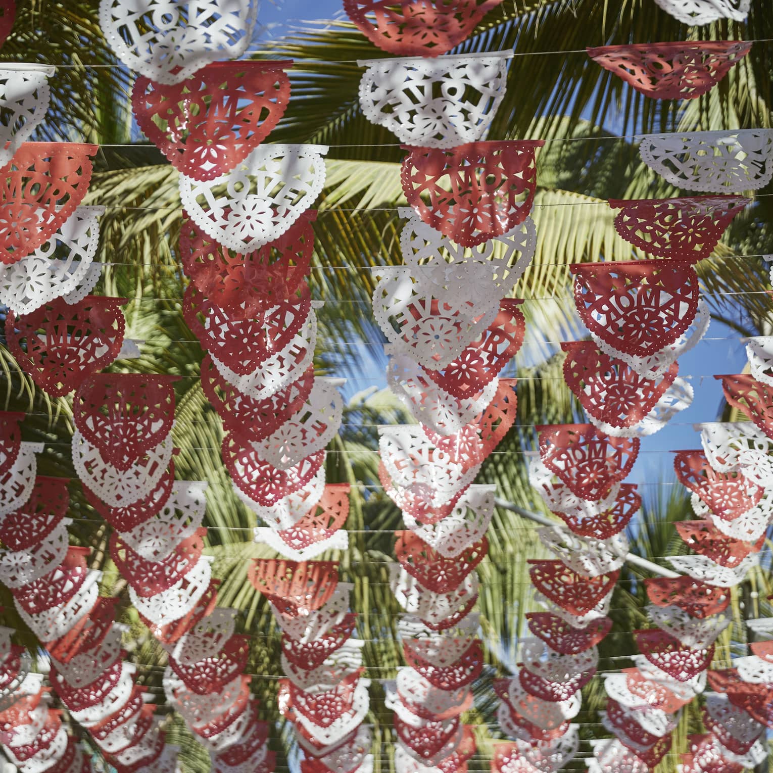 Looking up at rows of red and white Mexican Papel Picado banners hanging above, with palm trees and a sunny sky above them.