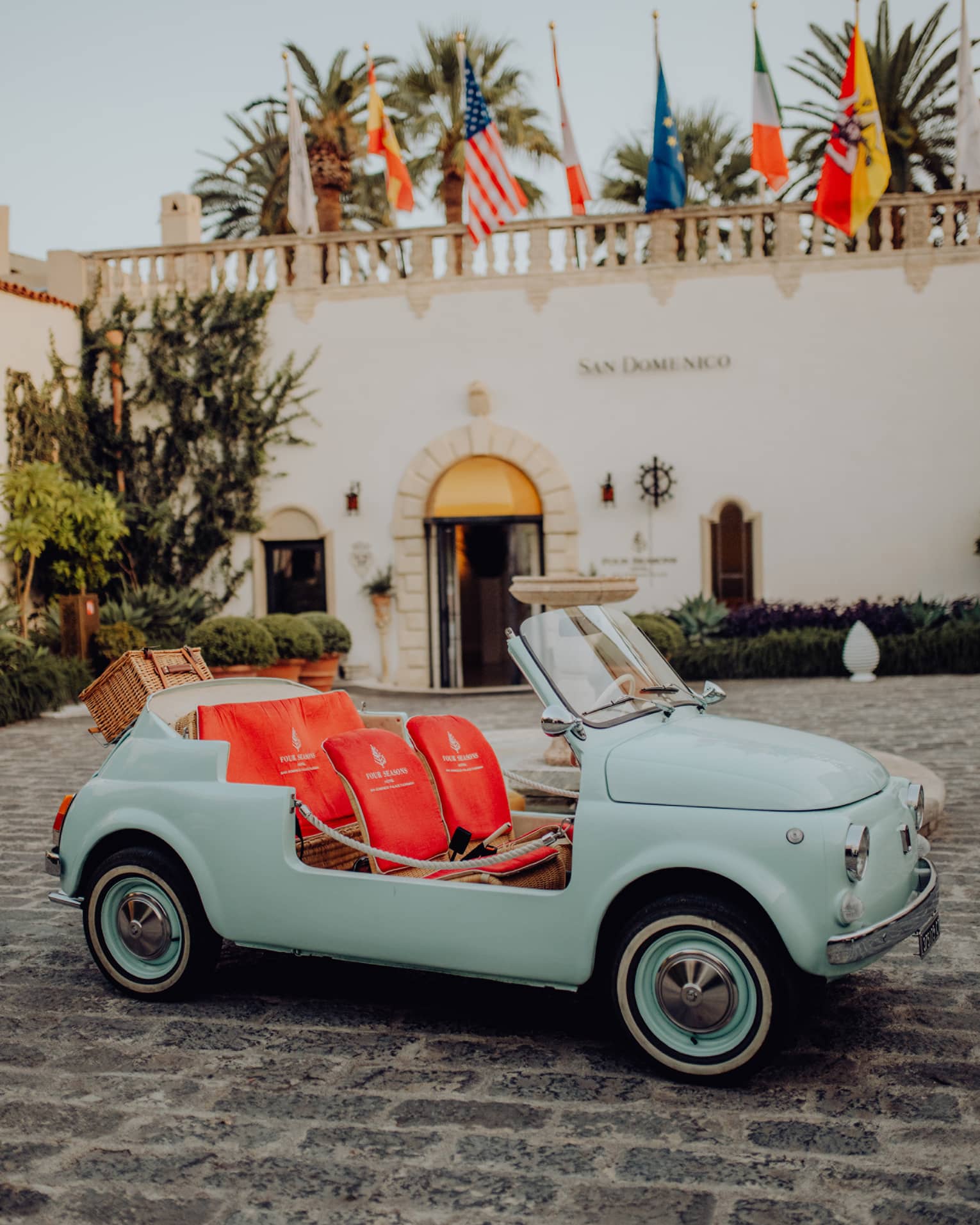 Vintage teal convertible Fiat 500 with bright coral seats parked on a cobblestone driveway in front of San Domenico Palace.