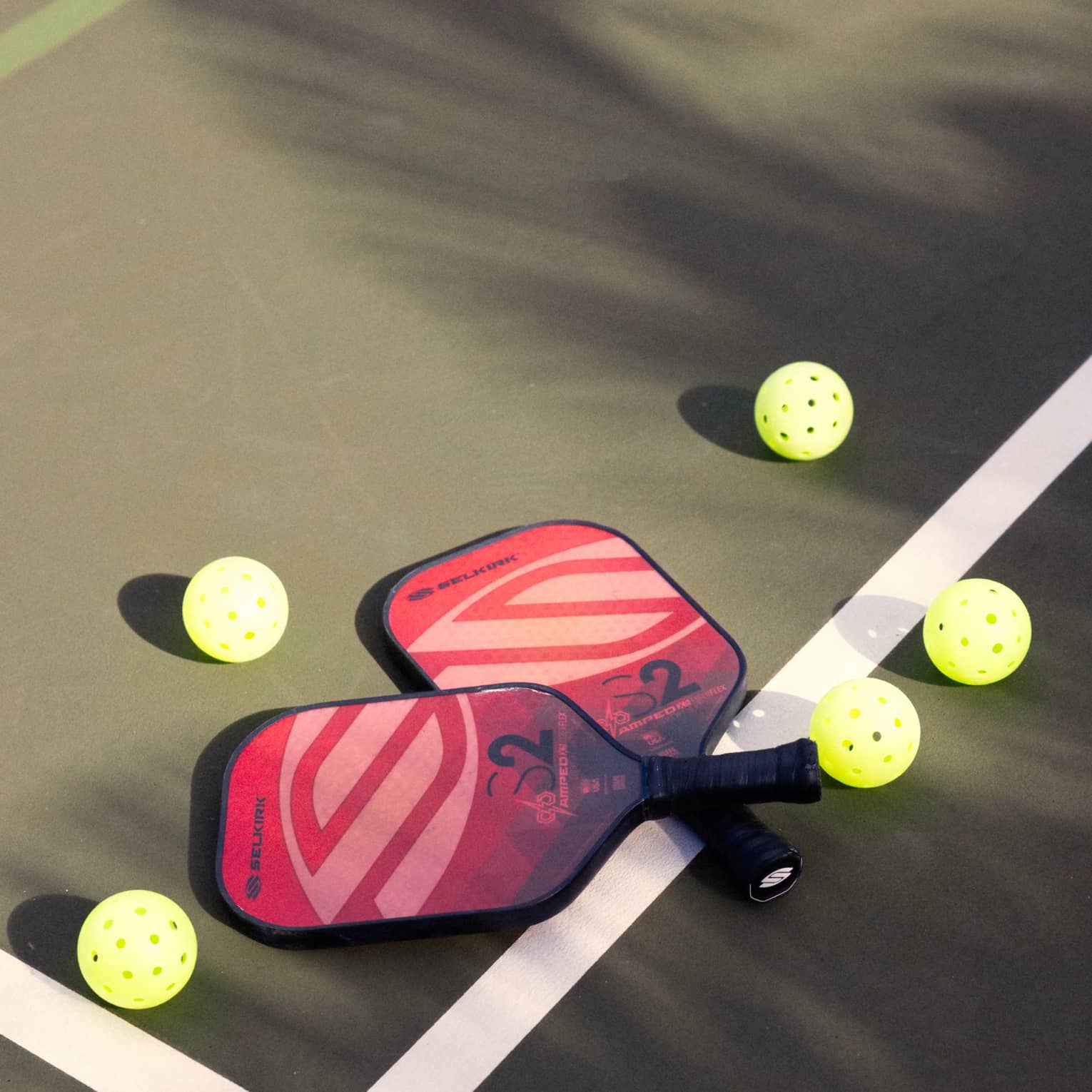 Close-up of two red and pink pickleball racquets, handles crisscrossed on a white court line, surrounded by five pickleballs.