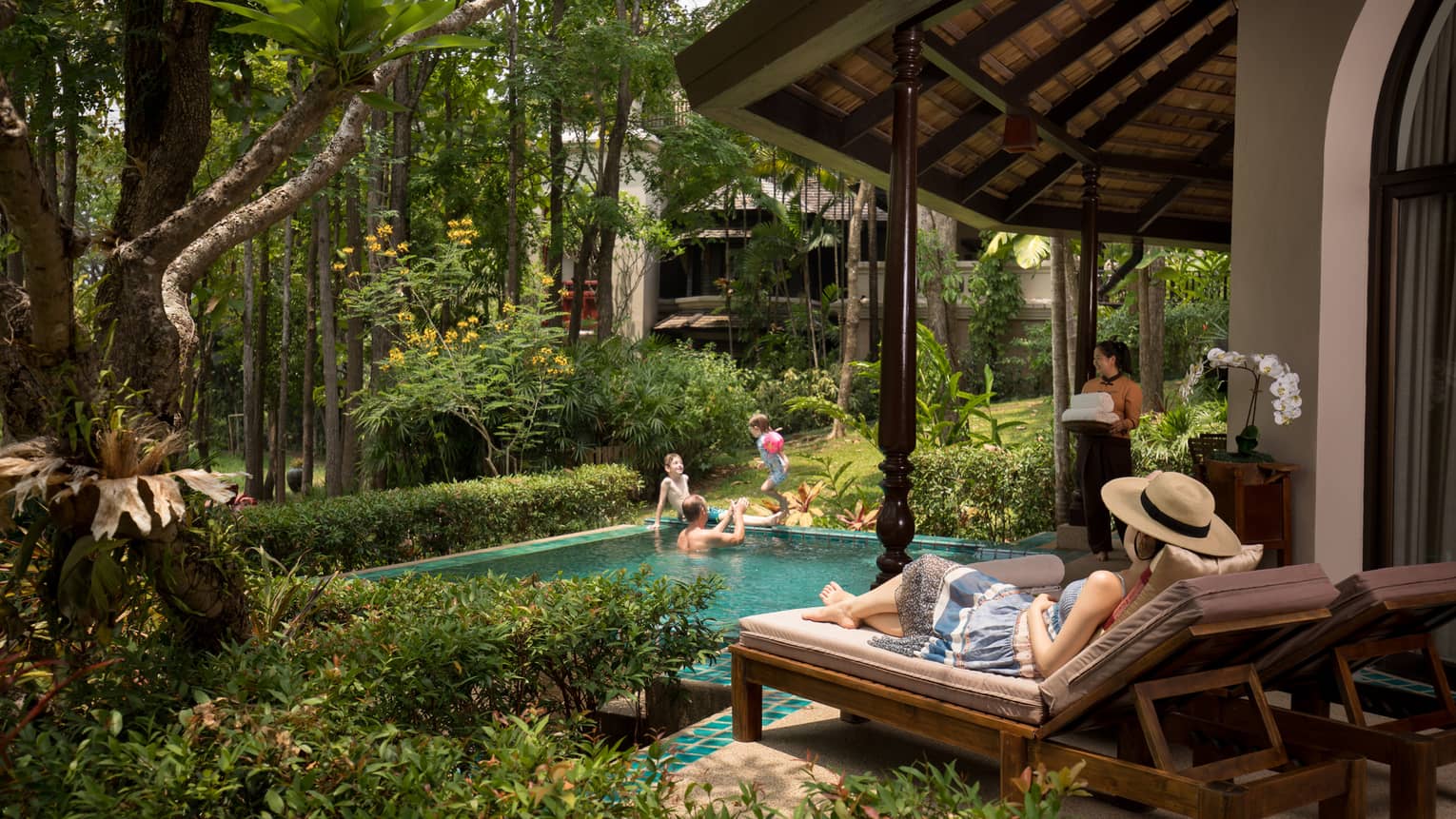 Person relaxing on a sun lounger beside a private pool while others play in the water and a staff member carries fresh towels nearby amidst lush greenery