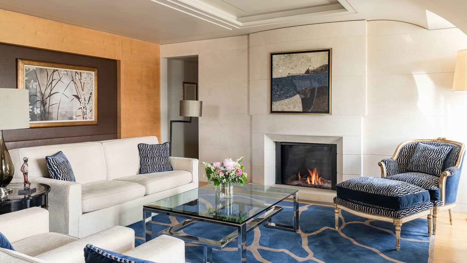 Luxury hotel suite living room with a cosy fireplace, cream sofa, blue and cream armchairs, glass coffee table, floral arrangement and wall art