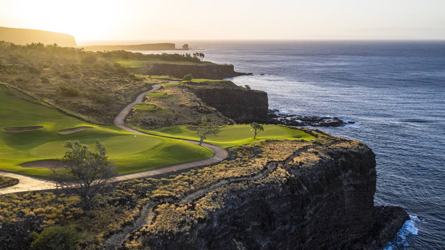 Soft sunlight filters over a pristine golf course with a winding road set on the edge of a cliff overlooking the calm ocean.
