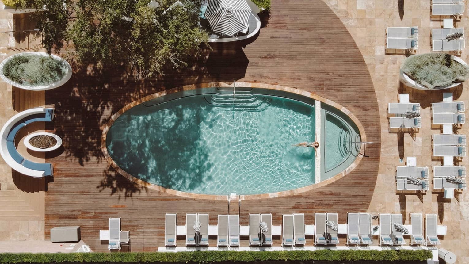 Aerial view of an oval pool surrounded by a rectangular wooden deck and rows of lounge chairs on two sides with greenery and more loungs seating on the other two sides