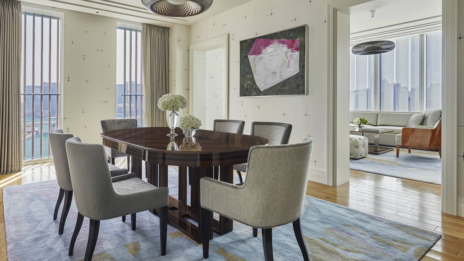 View of a stylish hotel room with a wooden cabinet, a purple chair at a desk, and large windows with gray curtains letting in natural light