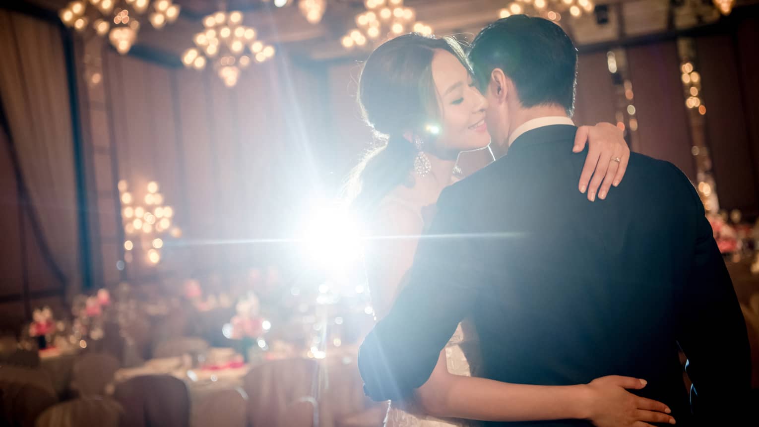 A bride and groom embrace each other in an indoor reception hall decorated with chandeliers and banquet tables