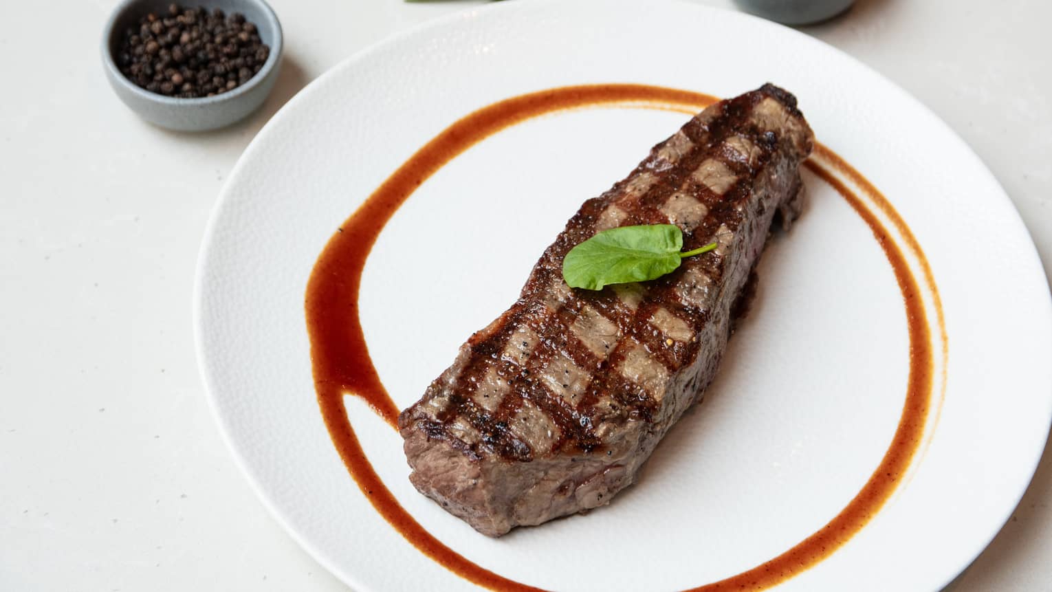 Strip steak topped with a green leaf for garnish and plated on a white round plate with a streak of sauce surrounding the steak
