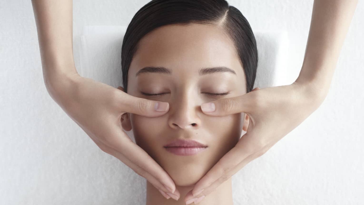 Woman in white bathrobe lies on massage table with eyes closed, masseuse rests hands on her cheeks