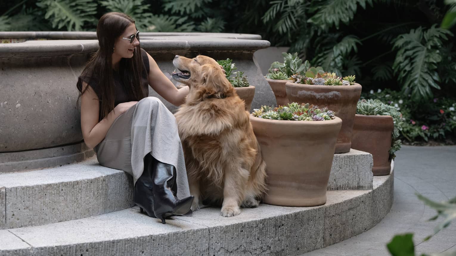 A guest and her dog sit on the steps surrounding an outdoor fountain. Greenery is in the background, and decorative potted plants are next to them.