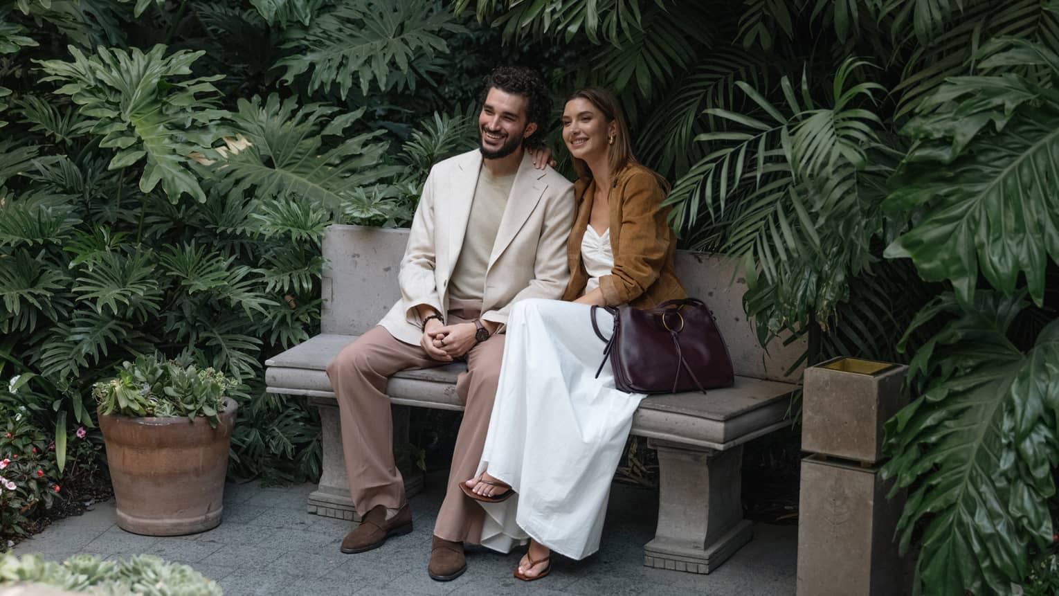 Two people sit on a bench outdoors, surrounded by greenery and plantings.