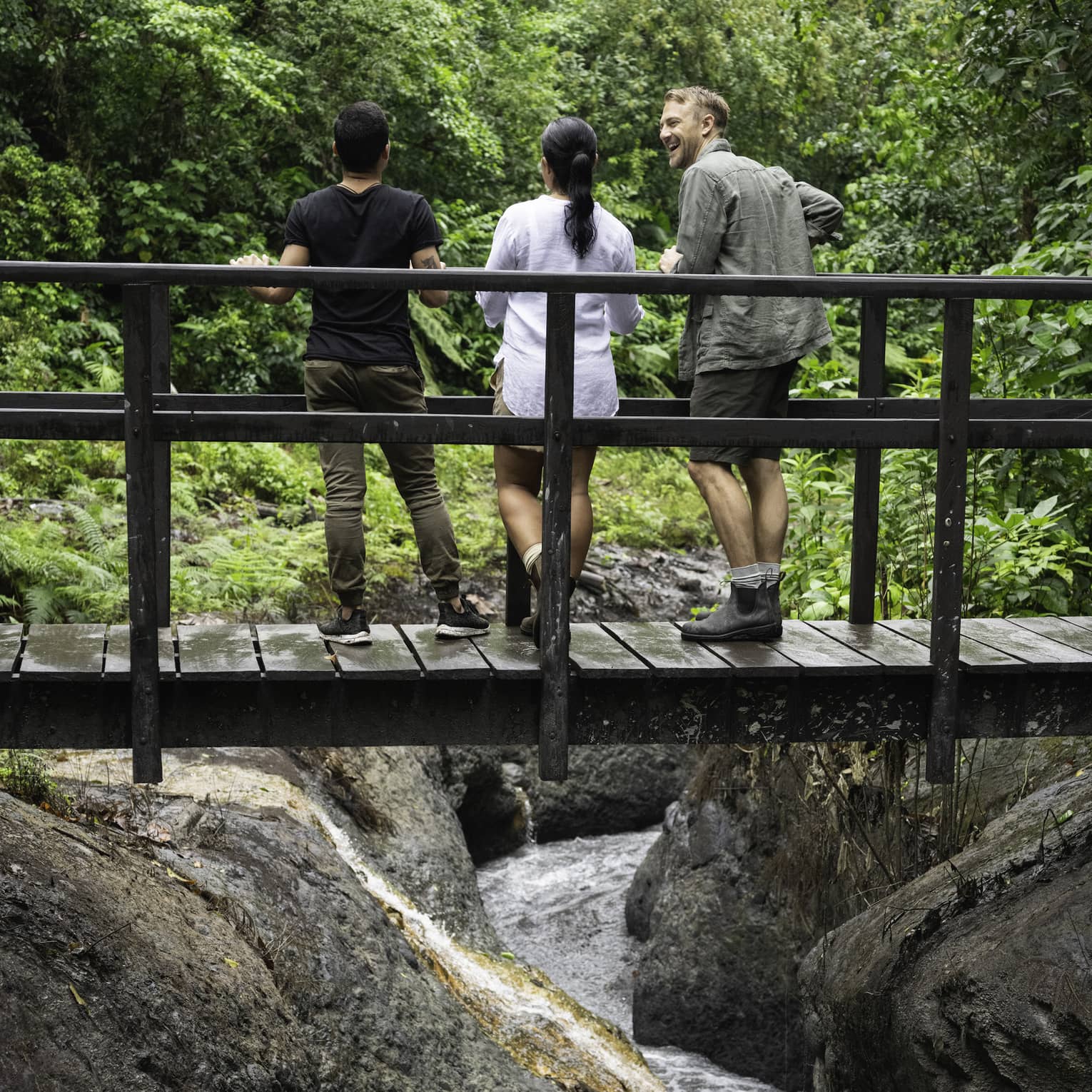 Three people stand on a wooden bridge that crosses over a small river in the middle of a forest