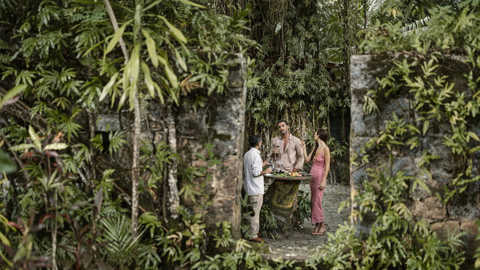 Framed by stone columns and surrounded by greenery are guests enjoying wine and cheese off a table made from a tree stump.